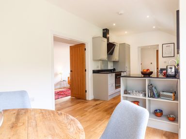 A kitchen with light grey cabinets a black countertop a wooden table grey chairs and an open door showing an adjacent room at Bluebell Cottage in Mary Tavy