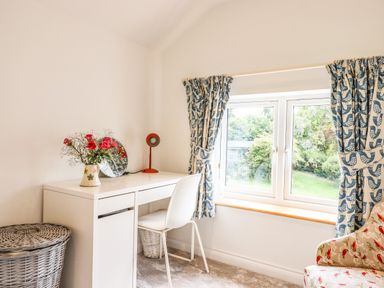 A small room with a white desk chair a vase with flowers on the desk a window with curtains and a patterned armchair at Bluebell Cottage Mary Tavy