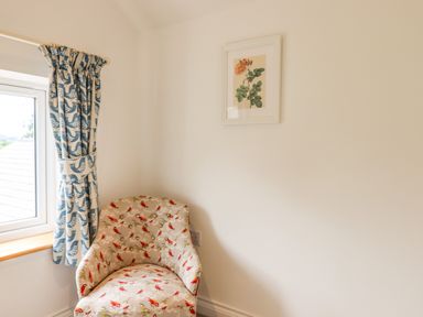 A corner of a room with a patterned armchair below a framed flower picture and a window with blue patterned curtains at Bluebell Cottage in Mary Tavy
