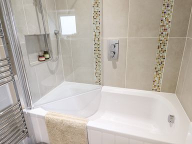 A bathroom with a white bathtub beige tiled walls and a towel hanging on the side of the tub at Bluebell Cottage in Mary Tavy