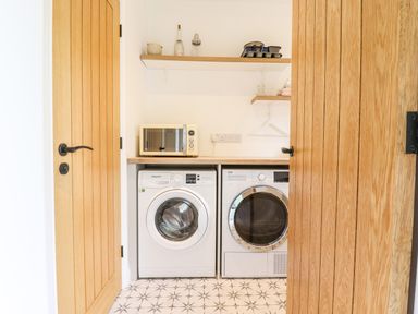 A laundry room with a washing machine dryer microwave and shelves with household items at Bluebell Cottage Mary Tavy