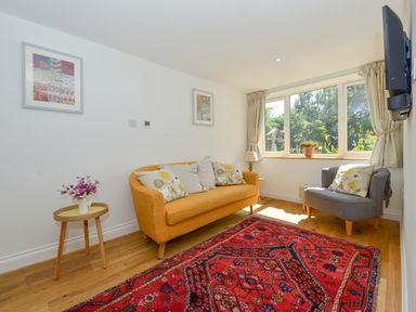 A sitting room with a yellow sofa a gray armchair a red patterned rug and windows with curtains at Bluebell Cottage in Mary Tavy