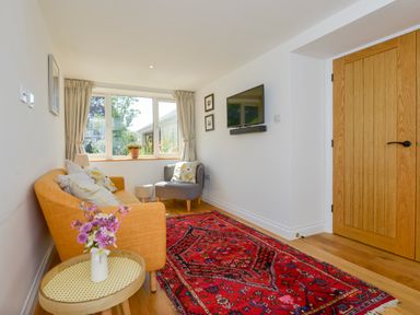 A living room with an orange sofa a gray armchair a red patterned rug a wooden door and a wall-mounted television at Bluebell Cottage in Mary Tavy