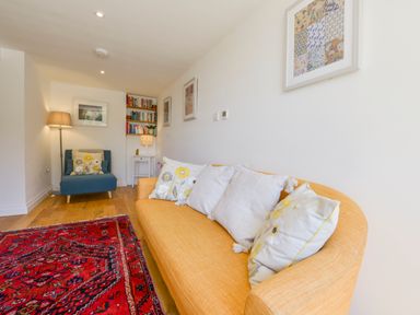 A living room with a yellow sofa with cushions a blue armchair a red patterned rug a floor lamp a small white table a bookshelf and framed pictures on the walls at Bluebell Cottage in Mary Tavy