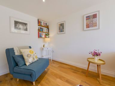 A blue chair with cushions near a small white side table with a lamp and wall shelves with books and a small wooden round table with flowers at Bluebell Cottage in Mary Tavy