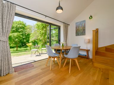 A dining area with a wooden table and four gray chairs next to large open glass doors leading to a garden at Bluebell Cottage in Mary Tavy