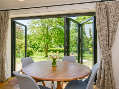 A dining area with a round wooden table and four grey chairs by open folding glass doors overlooking a garden at Bluebell Cottage in Mary Tavy