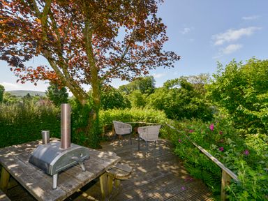 An outdoor wooden deck with a pizza oven on a table two chairs and surrounding trees and plants at Bluebell Cottage in Mary Tavy