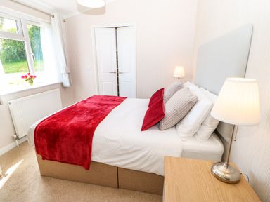 A bedroom with a bed featuring a red throw and pillows a bedside table with a lamp a window with curtains and flowers on the sill at Rashierigg in Catterick North Yorkshire