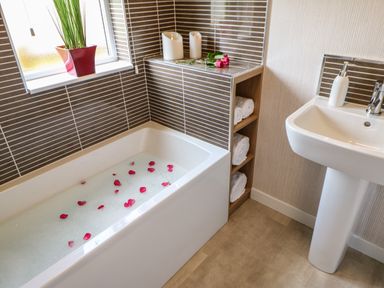 A bathroom with a bathtub filled with bubble bath and rose petals next to a sink with soap dispenser and shelves with rolled towels at Rashierigg in Catterick North Yorkshire