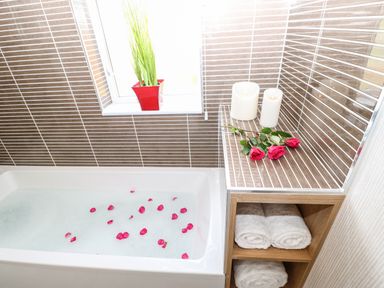 A bathroom with a bathtub filled with water and rose petals candles and red roses on a tiled surface and rolled white towels in wooden shelves at Rashierigg in Catterick North Yorkshire