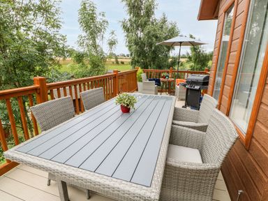 An outdoor wooden deck with a rectangular table, wicker chairs, a barbecue grill, and a small table with an umbrella at Rashierigg in Catterick North Yorkshire