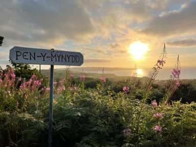 A landscape with pink flowers and a signpost reading Pen y Mynydd with a sunset over water in the background at Pen y Mynydd Newport Pembrokeshire