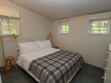 A bedroom with a bed covered by a patterned blanket two stools yellow lamps and windows at Pen y Mynydd in Newport Pembrokeshire