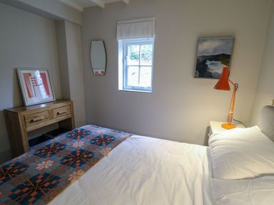 A bedroom with a bed with white sheets a wooden dresser with framed art an orange lamp and a window at Pen y Mynydd Newport Pembrokeshire
