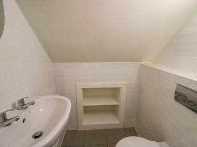 A small bathroom with a white sink and dual faucets a wall shelf under a slanted ceiling and a toilet at Pen y Mynydd in Newport Pembrokeshire