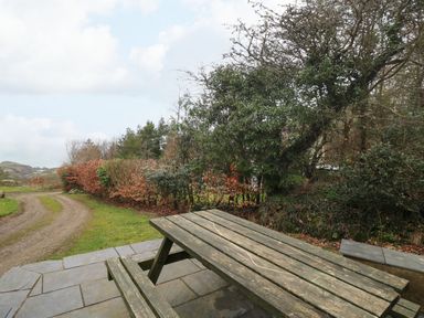 A wooden picnic table on a stone patio near a dirt path and trees at Pen y Mynydd in Newport Pembrokeshire