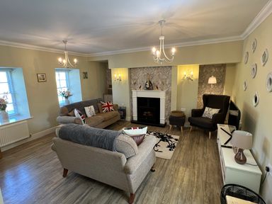 A living room with two sofas and an armchair around a fireplace with wall sconces and chandeliers at May Bell Cottage in Dersingham