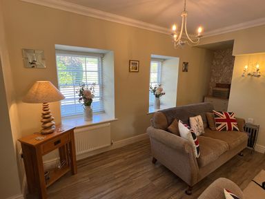 A living room with a brown sofa and pillows next to windows with white blinds and flowers on the sills at May Bell Cottage in Dersingham