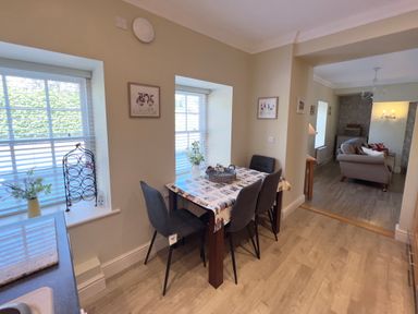 A dining area with a table and four chairs next to two windows and a living room in the background at May Bell Cottage in Dersingham