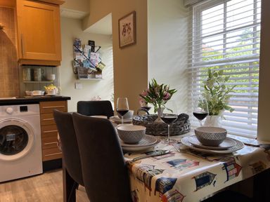A kitchen dining area with a table set with plates bowls wine glasses and flowers next to a window with blinds at May Bell Cottage in Dersingham