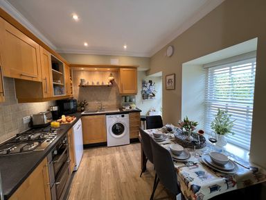 A kitchen with wooden cabinets a black countertop a dining table set with dishes glasses and flowers at May Bell Cottage in Dersingham