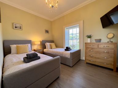 A bedroom with two single beds a wooden dresser a wall-mounted TV and a window with blinds at May Bell Cottage in Dersingham