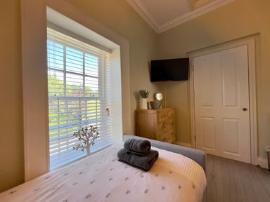 A bedroom with a bed near a window and a wooden dresser with a TV mounted above at May Bell Cottage in Dersingham