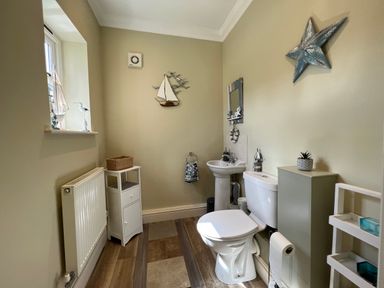 A bathroom with a toilet, sink, storage units, wall decorations, and a window at May Bell Cottage in Dersingham