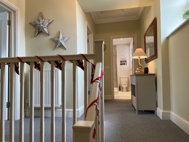 A hallway with a railing, two star-shaped wall decorations, a cabinet with a lamp and a mirror, and a bathroom visible at the end at May Bell Cottage in Dersingham