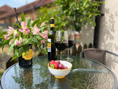 A glass table with a bottle used as a vase with pink flowers a wine bottle two glasses of red wine and a bowl of fruit on a patio at May Bell Cottage in Dersingham