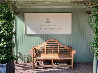 A wooden bench against a green wooden wall with a welcome to Sandringham sign and plants on the sides