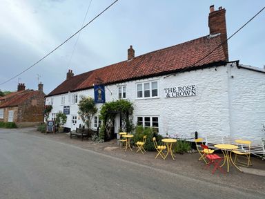 A white building with a red tiled roof and outdoor seating with yellow and red tables and chairs at The Rose and Crown