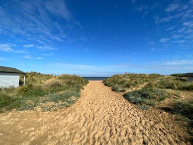 A sandy path leading between grass-covered dunes to a beach with the sea and blue sky in the background at May Bell Cottage in Dersingham
