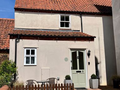 Exterior of a small house with a green door marked number 5 two windows and a small fenced patio with table and chairs at May Bell Cottage in Dersingham