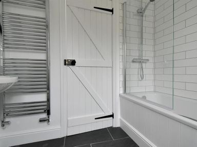 A bathroom with a white wooden door a bathtub with a glass shower screen and a wall-mounted showerhead and a chrome towel radiator at 12 Mill Green in Lyme Regis