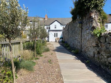 A paved pathway between a stone wall and wooden fence leading to a white house with garden plants and trees at 12 Mill Green in Lyme Regis