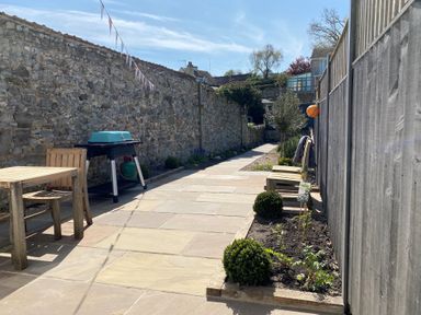 An outdoor patio with a stone wall and wooden furniture including a table, chairs, and benches with a barbecue and small garden beds at 12 Mill Green in Lyme Regis