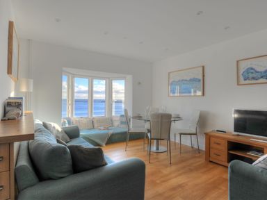 A living room with blue sofas a dining table and chairs wooden floor and a bay window with sea view at Sea Fever in Lyme Regis