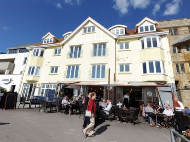People dining outside a restaurant in front of a three-story building with white walls and multiple windows at Sea Fever in Lyme Regis
