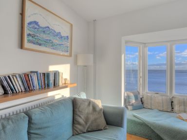 A living room with a blue sofa a shelf of books a floor lamp and a window seat with cushions overlooking the sea at Sea Fever in Lyme Regis