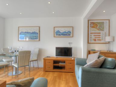 A living room with blue armchairs a wooden TV stand glass dining table with chairs and framed pictures at Sea Fever Lyme Regis