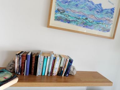 A wooden shelf with books supported by two rocks and a framed abstract blue artwork on a white wall at Sea Fever in Lyme Regis