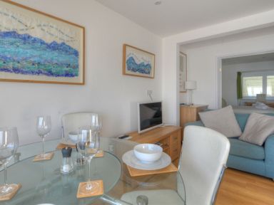 A dining area with a glass table set with bowls and wine glasses next to a living room with a blue sofa and two framed artworks on the wall at Sea Fever in Lyme Regis