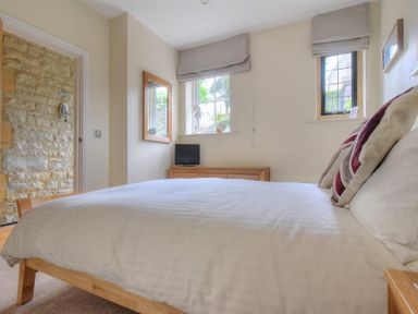 A bedroom with a wooden bed frame and white bedding a wooden dresser with a small black tv two windows with gray blinds and a stone wall visible through a doorway at Coram Lodge in Lyme Regis