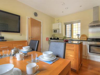 A kitchen with a wooden dining table set with cups bowls and glasses near an island and stainless steel oven at Coram Lodge in Lyme Regis