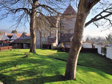 A grassy garden with two leafless trees and a stone building in the background at Coram Lodge in Lyme Regis