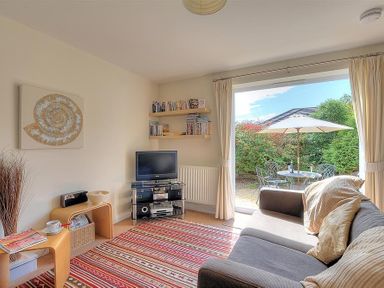 A living room with a sofa a tv on a stand shelves with books and an open door leading to a patio with a table umbrella and chairs at 1 Coppers Knapp in Lyme Regis