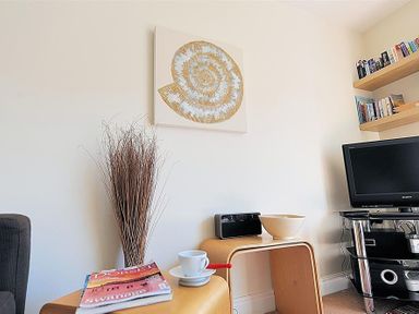 A corner of a living room with a TV on a stand shelves with books a small table with a radio a bowl and a cup a magazine and a bundle of dried sticks on the floor at 1 Coppers Knapp in Lyme Regis