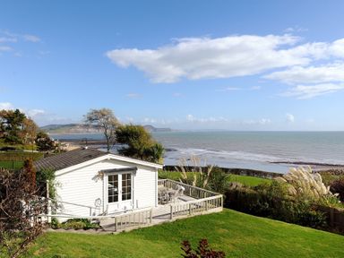 A small white cabin with a deck overlooking the sea and a grassy garden at The Cabin in Lyme Regis